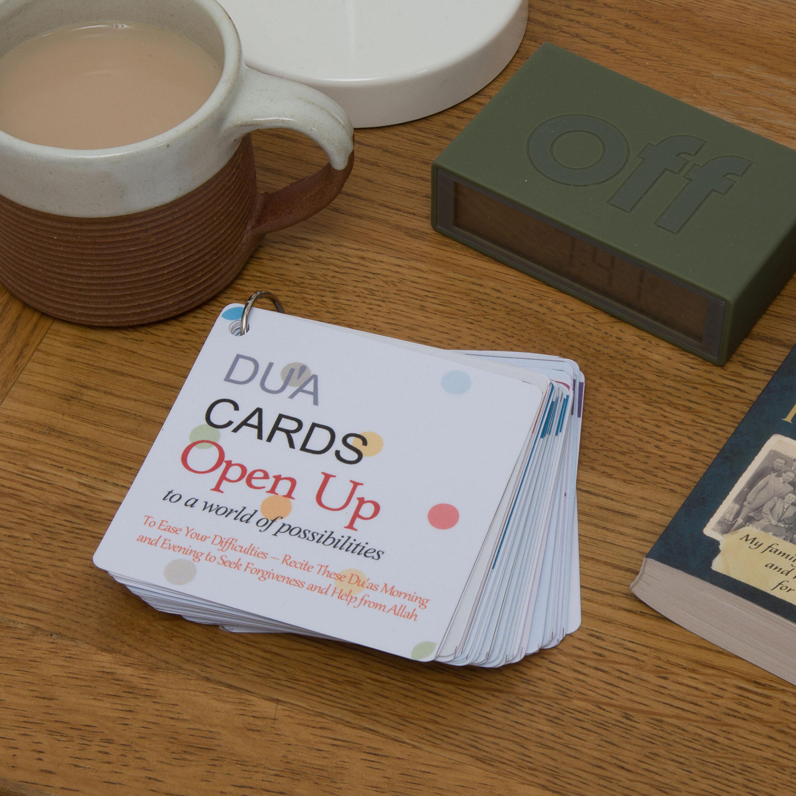 Stack of Dua Cards with text 'Open Up to a world of possibilities' on a table with a cup of tea, a green 'off' timer, and a vintage book in the background, suggesting a peaceful reading or meditation space.